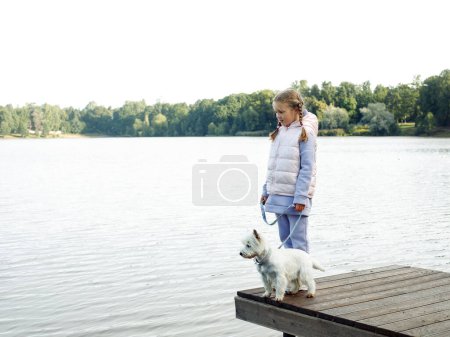 Téléchargez les photos : A seven-year-old girl with a small white dog stands on a bridge near the lake and looks at the water. Friendship with pets. - en image libre de droit