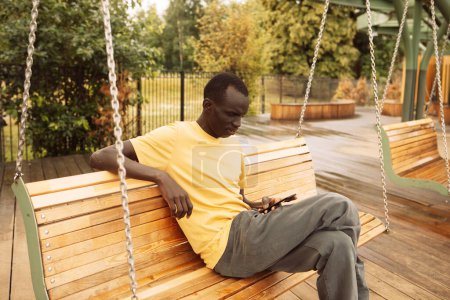 Photo for Dark-skinned african american young man sitting on a swing in the park on a summer day, holding a smartphone and texting or browsing the news on social networks. - Royalty Free Image
