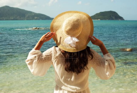 Foto de Joven morena en vestido blanco de verano con sombrero de paja mirando al cielo y al mar. Vista trasera. - Imagen libre de derechos
