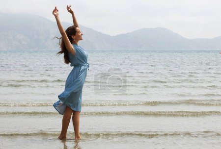 Photo for Young brunette woman in a blue dress walking barefoot on a beach and dangles his feet in the water. Happy summer vacation. - Royalty Free Image