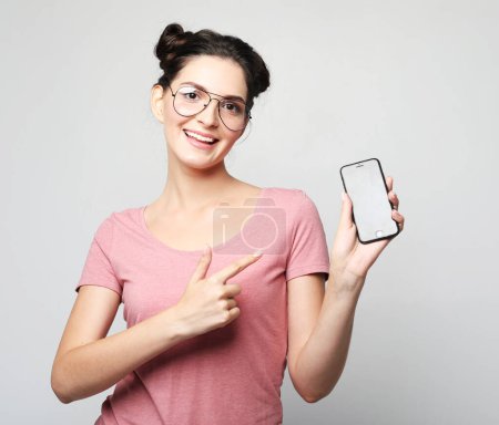 Téléchargez les photos : Portrait d'une jeune femme brune souriante portant des lunettes de vue montrant ou présentant un téléphone portable sur fond gris. - en image libre de droit