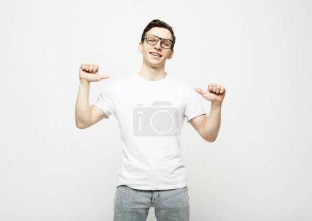 Photo for Happy young man in a white t-shirt and glasses shows himself, thumbs up. Joy, pleasure, self-presentation. Lifestyle concept. - Royalty Free Image