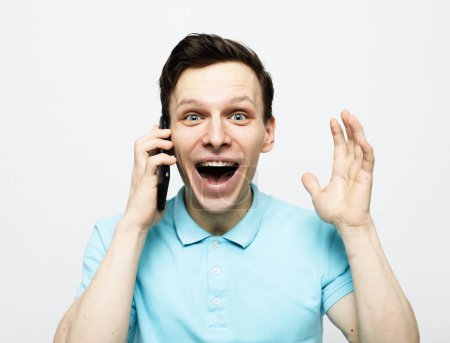 Photo for Enthusiastic young man talks via mobile phone, got call from friend or colleague, tells something emotionally, discusses good news, raises hand, holds cellular near ear, wears blue clothes. - Royalty Free Image