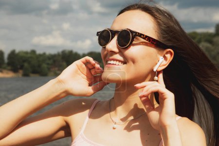 Téléchargez les photos : Joyeux jeune femme heureuse avec les cheveux longs dans les lunettes de soleil en écoutant de la musique à côté du lac. Bon été. Concept de style de vie. - en image libre de droit