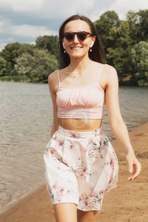 Foto de Una hermosa joven vestida con ropa casual camina por la playa cerca del lago. Feliz verano.. - Imagen libre de derechos
