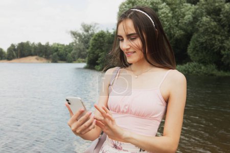 Téléchargez les photos : Jeune femme souriante dans occasionnel près du lac utiliser téléphone portable chat en ligne sur le réseau social. L'heure d'été. Concept de mode de vie actif. - en image libre de droit
