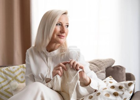 Photo for Portrait of smiling pleasant old mature blond woman sitting on sofa with handmade clothes in hands, enjoying knitting warm scarf with woolen needles, retired people leisure hobby activity. - Royalty Free Image