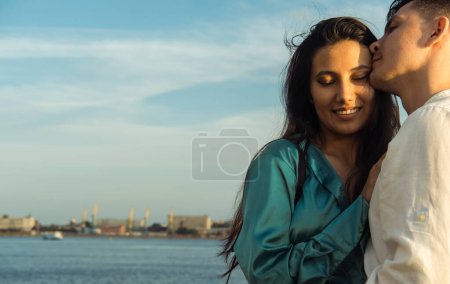 A young loving couple of different nationalities admires the sunset near the water on the embankment. Asian woman and European man. Happy summer time.