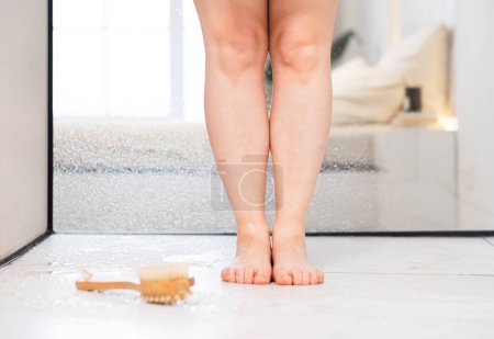 Téléchargez les photos : Les jambes des femmes se rapprochent sous la douche. A proximité se trouve une brosse pour le massage anti-cellulite. Le concept d'autosoin et de santé. - en image libre de droit