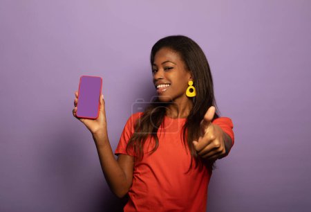 Foto de Retrato de positiva alegre afroamericana promotor show smartphone, presente nuevo dispositivo moderno desgaste naranja camiseta aislada sobre fondo de color púrpura - Imagen libre de derechos