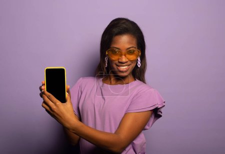 Foto de Joven mujer afroamericana sonriente con el pelo largo aislado sobre fondo morado, vistiendo una camiseta lila y gafas de sol, sosteniendo el teléfono en blanco y mostrando una oferta interesante. - Imagen libre de derechos