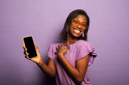 Foto de Joven mujer afroamericana sonriente con el pelo largo aislado sobre fondo morado, vistiendo una camiseta lila y gafas de sol, sosteniendo el teléfono en blanco y mostrando una oferta interesante. - Imagen libre de derechos