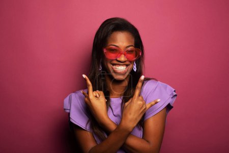 Foto de Joven dama negra con blusa lila y gafas de sol rosadas apuntando hacia arriba sobre fondo rosa del estudio. Millennial mujer afroamericana teniendo idea creativa. - Imagen libre de derechos