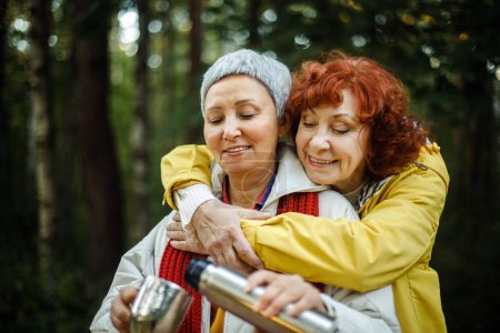 Téléchargez les photos : Two elderly woman friends walk in the forest, pour coffee from a thermos, have a great time together, an elderly happy couple. Lifestyle and golden age concept. - en image libre de droit