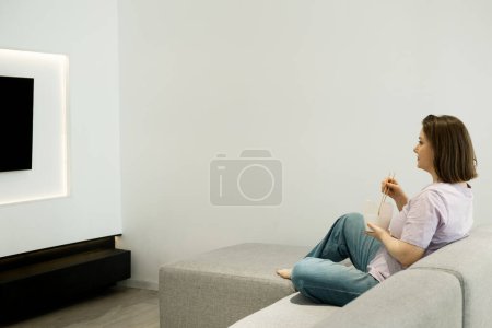 Photo for Young woman eating noodles with chopsticks, holding paper box at home in living room. Woman sitting on sofa and watching tv. Food delivery, fastfood concept. - Royalty Free Image