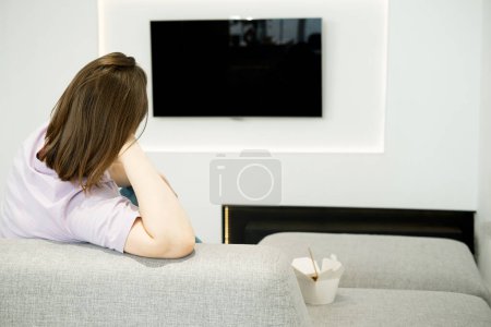 Téléchargez les photos : A young woman is sitting on a sofa, watching TV, Asian food in a box is nearby, a lonely evening. - en image libre de droit