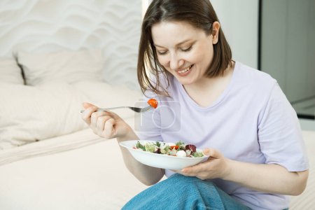 Photo for Pretty adult oversized woman sitting on bed and eating fresh vegetable salad - Royalty Free Image