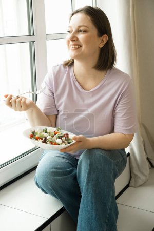 Foto de Mujer feliz de gran tamaño junto a la ventana y comer ensalada de verduras frescas. Concepto de estilo de vida. - Imagen libre de derechos