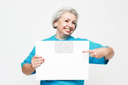 Téléchargez les photos : Les personnes âgées et le concept de style de vie : Une femme âgée heureuse et charmante tient une feuille de papier vierge dans ses mains et la pointe du doigt. - en image libre de droit