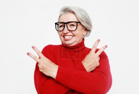 Foto de Retrato de una divertida mujer de pelo gris con gafas y suéter rojo haciendo un símbolo de rock and roll sobre fondo blanco. Concepto de estilo de vida. - Imagen libre de derechos