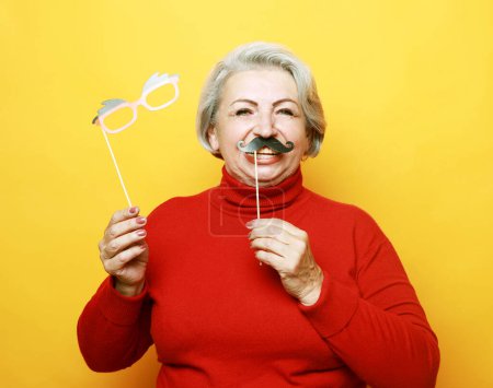 Photo for Lifestyle and old people concept: funny grandmother in red sweater with fake mustache and glasses, laughs and prepares for party over yellow background - Royalty Free Image