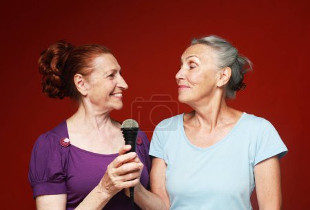 Téléchargez les photos : Lifestyle, friendship and old people concept. two elderly female friends with microphone, laughs and prepares for party karaoke over red background - en image libre de droit