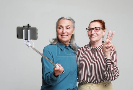 Foto de Estilo de vida, amistad, tehnología y concepto de personas mayores. Dos encantadoras amigas ancianas haciendo selfie sobre fondo gris. - Imagen libre de derechos