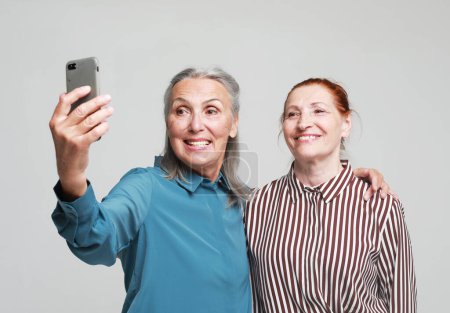 Lifestyle, friendship, tehnology and old people concept. Two charming elderly women friends making selfie over grey background.