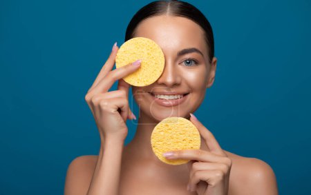 Photo for Young female model holding yellow sponges near her face. Portrait of beautiful woman with clean face on blue color background. - Royalty Free Image