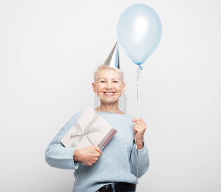 Foto de Concepto de personas, envejecimiento y madurez. Mujer anciana asombrada con pelo corto con sombrero de fiesta sosteniendo globo azul y caja de regalo sobre fondo gris blanco. Mirando la cámara. - Imagen libre de derechos