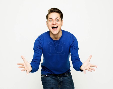 Foto de Estilo de vida y concepto de emoción: Feliz joven guapo con suéter azul haciendo gestos y manteniendo la boca abierta sobre fondo blanco - Imagen libre de derechos