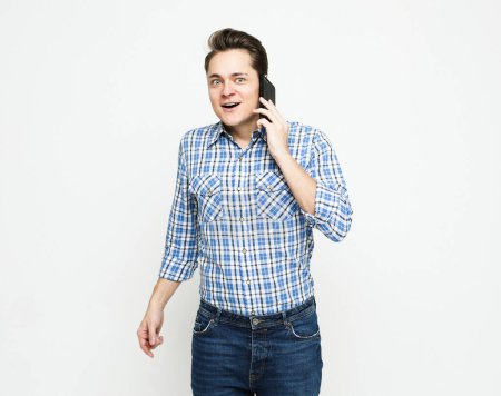 Foto de Joven hombre guapo con camisa a cuadros azul haciendo gestos y sonriendo mientras habla en el teléfono móvil sobre fondo blanco - Imagen libre de derechos