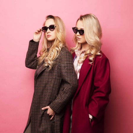 Foto de Moda dos mujeres rubias en abrigo con gafas de sol posando sobre fondo rosa. Moda otoño invierno foto. Dos hermanas.. - Imagen libre de derechos