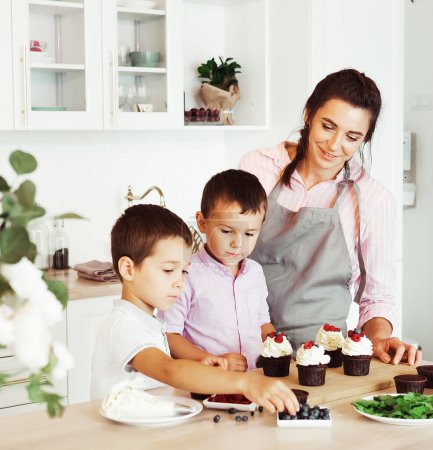 Photo for Happy family in the kitchen. Young Mom and two little boys, brothers make homemade cupcakes with berries. Family creativity, hobbies, joint activities with children. - Royalty Free Image