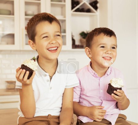Téléchargez les photos : Funny two brothers male kids are sitting together at the kitchen table and eating cupcakes. Lifestyle concept. - en image libre de droit