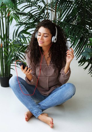 Téléchargez les photos : Belle jeune femme afro-américaine habillée occasionnellement écouter de la musique et assis sur le sol à la maison. Concept de style de vie. - en image libre de droit