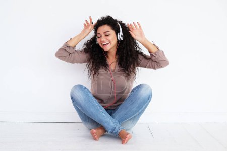 Téléchargez les photos : Jeune femme afro américaine heureuse portant des écouteurs assis sur le sol et écoutant de la musique, excité mixte race féminine - en image libre de droit