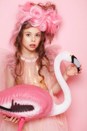Téléchargez les photos : Belle petite fille en robe rose avec un maquillage lumineux tenant un oiseau flamant rose sur fond rose - en image libre de droit