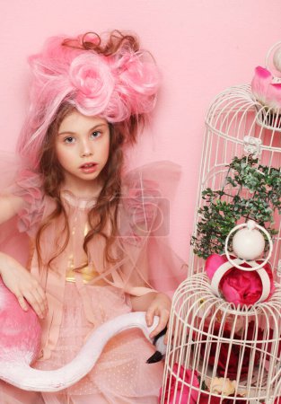 Foto de Hermosa niña en vestido rosa con maquillaje brillante sosteniendo un pájaro flamenco sobre fondo rosa - Imagen libre de derechos
