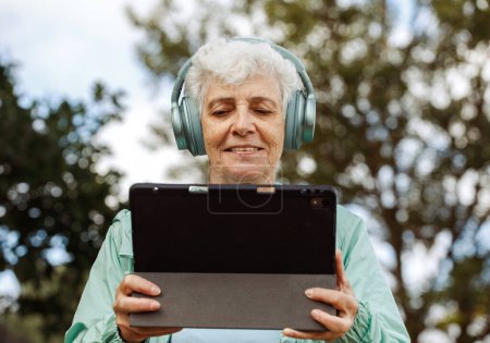Photo for Grandmother in headphones with a tablet listens to music. An elderly woman in headphones with a tablet sits on a park bench and listens to music. - Royalty Free Image