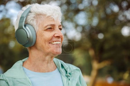 Téléchargez les photos : Vieille femme dans les écouteurs écoute de la musique dans le parc, concepts sur les personnes âgées, l'ancienneté et le vieillissement du bien-être - en image libre de droit