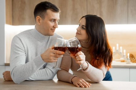 Téléchargez les photos : Romantic beautiful young couple holding glasses standing in modern cozy kitchen room interior, happy married guy husband and girl wife drinking red wine celebrate together at home - en image libre de droit