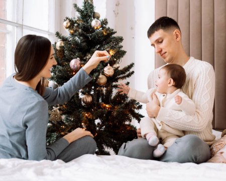 Téléchargez les photos : Joyeux jeune famille - maman et papa avec la petite fille à côté de l'arbre de Noël, amusez-vous, profitez de la vie familiale heureuse. Concept de style de vie. - en image libre de droit