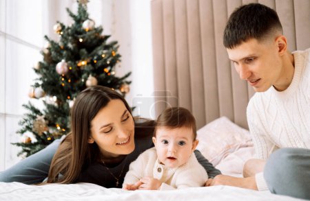 Téléchargez les photos : Joyeux jeune famille - maman et papa avec la petite fille à côté de l'arbre de Noël, amusez-vous, profitez de la vie familiale heureuse. Concept de style de vie. - en image libre de droit