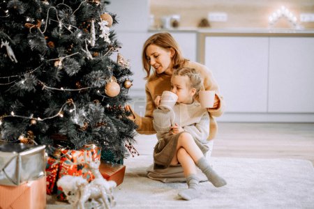 Téléchargez les photos : Concept de famille, vacances d'hiver et personnes - mère heureuse et petite fille décorant l'arbre de Noël à la maison - en image libre de droit
