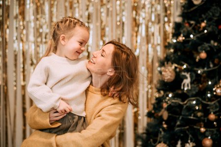 Foto de Una joven madre sostiene a una hija riendo en sus brazos junto al árbol de Navidad. Familia, vacaciones de invierno y concepto de personas. - Imagen libre de derechos
