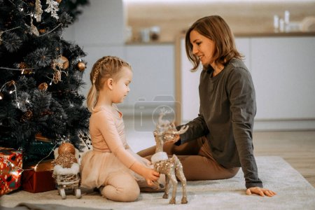 Téléchargez les photos : A young mother and little daughter play sitting near the Christmas tree. Family, winter holidays and people concept. - en image libre de droit