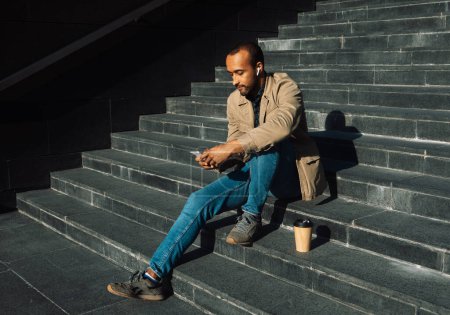 Photo for Portrait of young african american man wearing sunglasses sitting on steps using mobile - Royalty Free Image
