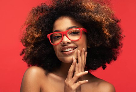 Téléchargez les photos : Happy Afro woman wearing eyeglasses smiling, has good mood isolated on studio red background. Excited african american female. Positive life concept. - en image libre de droit