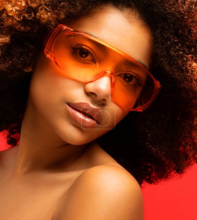 Téléchargez les photos : Young beautiful african american woman wearing orange sunglasses. Glamour makeup. Portrait over Red Background. - en image libre de droit